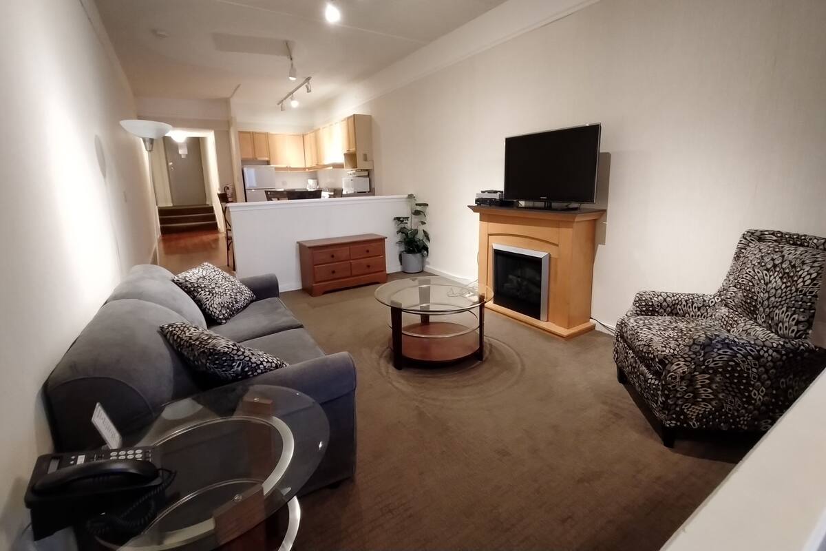 A living room with a grey sofa, patterned armchair, and a glass coffee table. A flat-screen TV is mounted above a fireplace.