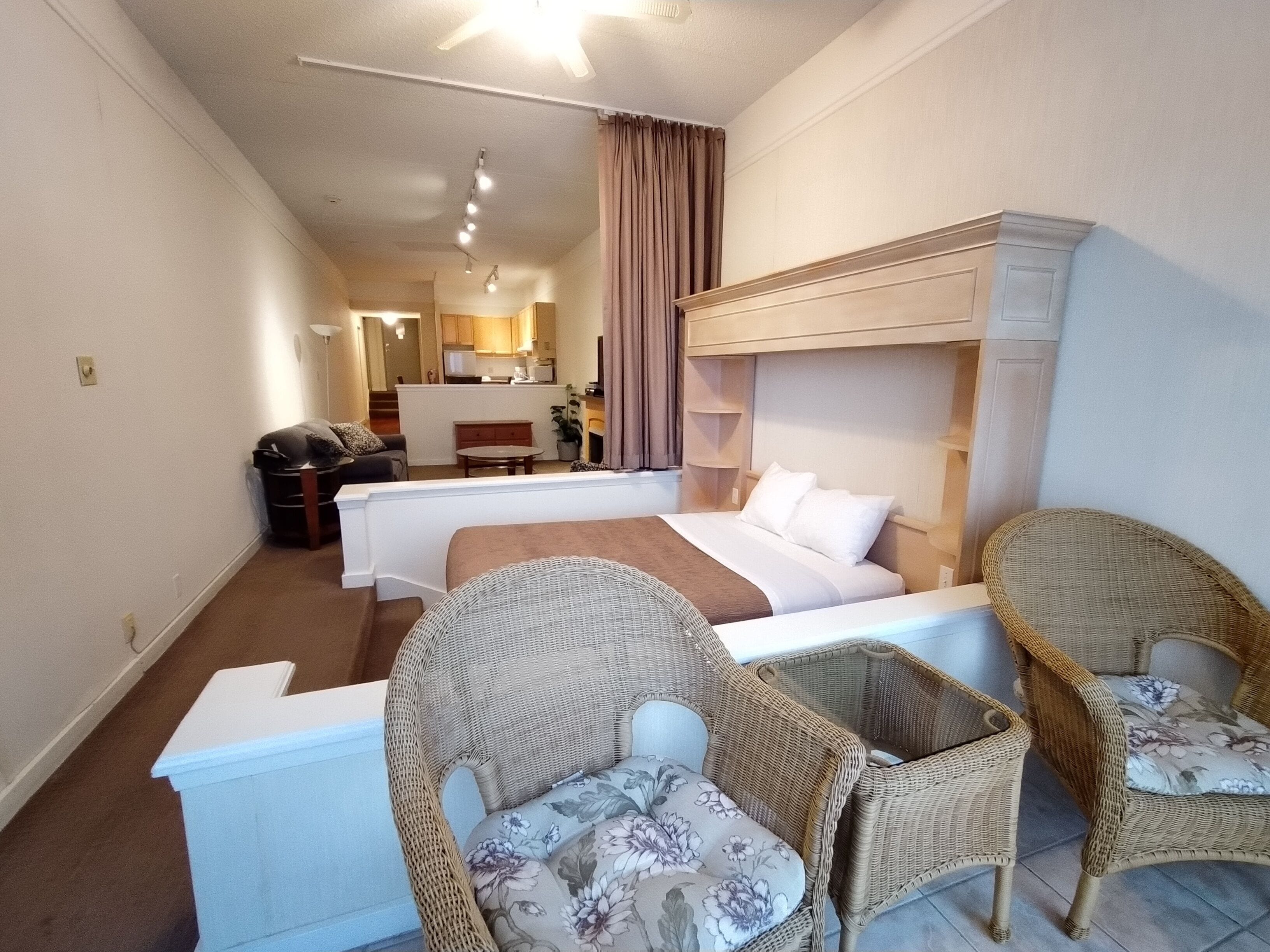 Une chambre d’hôtel comprenant un lit, deux fauteuils en osier, une petite table, un bureau, une kitchenette et une salle de bain.