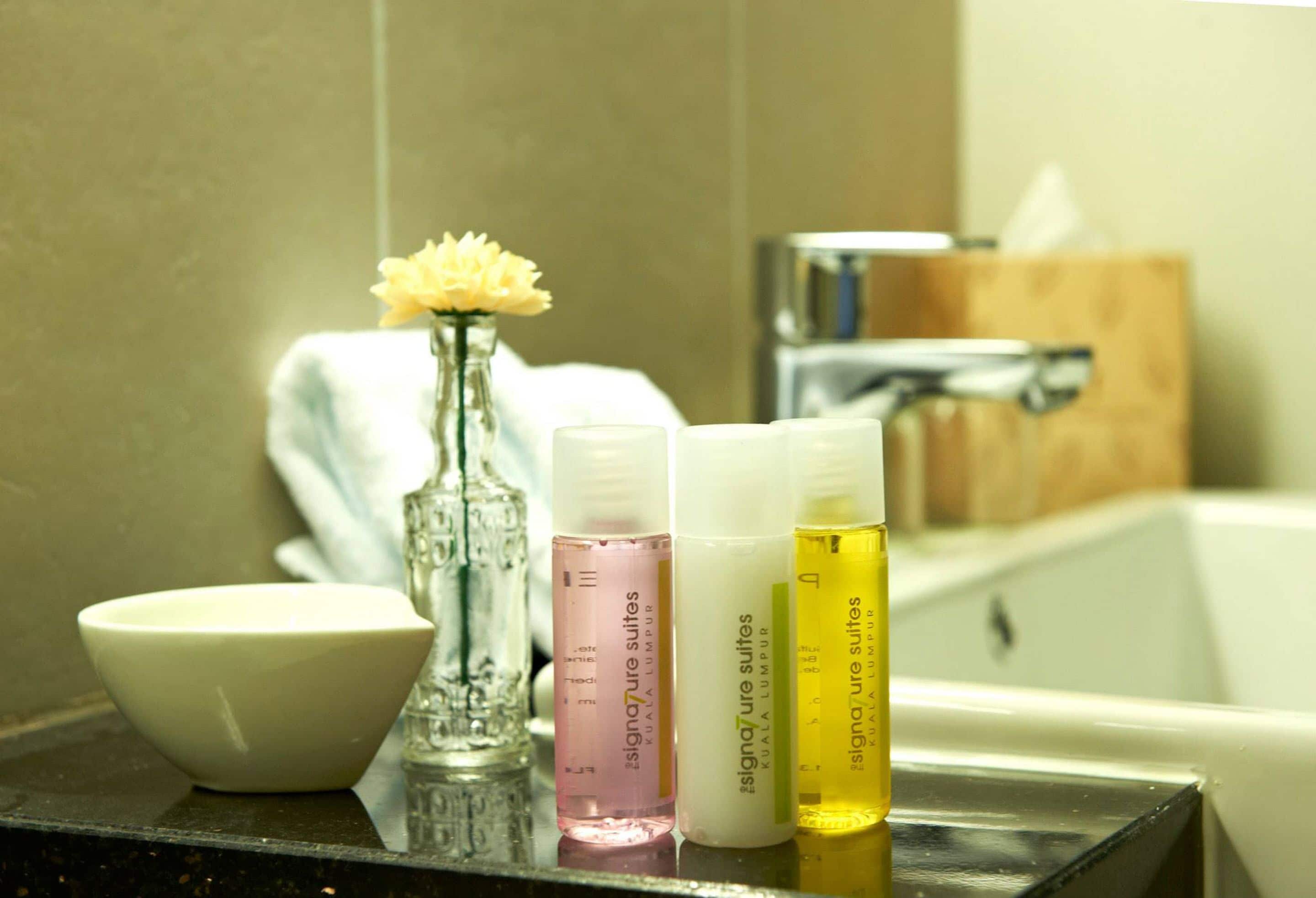 A bathroom counter with a white bowl, a glass vase with yellow flowers, and three bottles of Signature Suites shampoo.