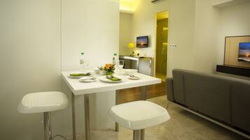 A modern hotel room with a dining area featuring a white table, two white stools, and a grey sofa. There is a painting on the wall and an air conditioning unit above the door.