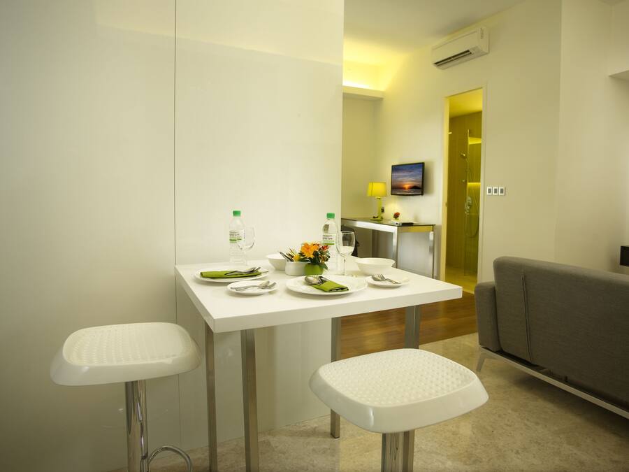 A modern hotel room with a dining area featuring a white table, two white stools, and a grey sofa. There is a painting on the wall and an air conditioning unit above the door.