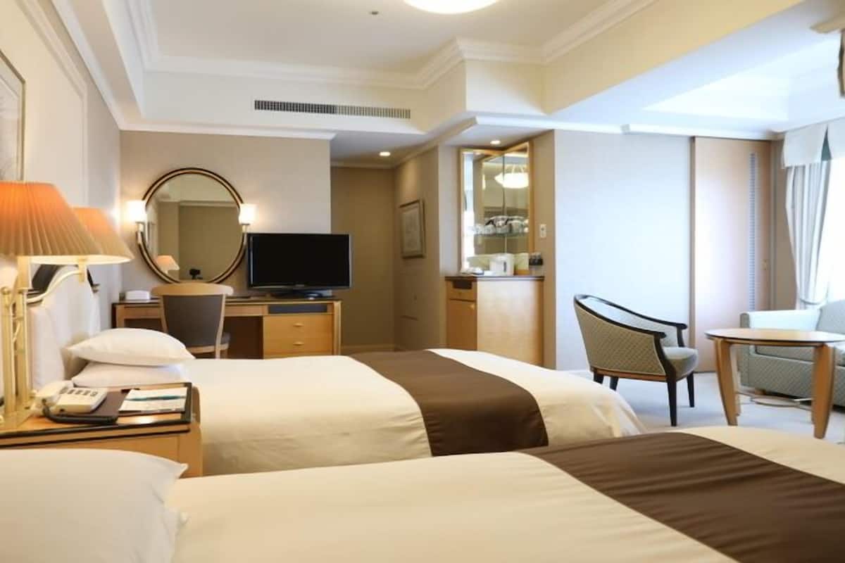 A hotel room with two beds, a desk with a TV, a chair, and a small table with a lamp.