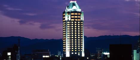 Un edificio de gran altura con numerosas ventanas iluminadas, rodeado de otros edificios y una cordillera al fondo.