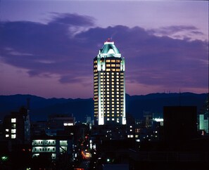 A high-rise building with multiple illuminated windows, surrounded by other buildings and a mountain range in the background. - Imabari Kokusai Hotel (Imabari)