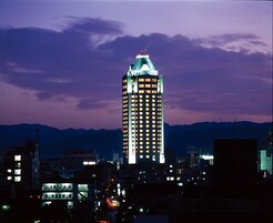 Un edificio de gran altura con numerosas ventanas iluminadas, rodeado de otros edificios y una cordillera al fondo.
