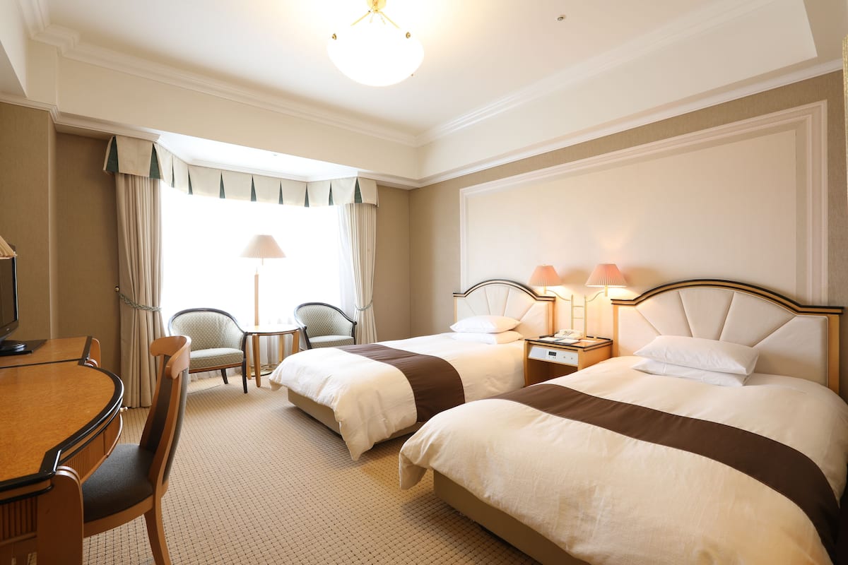 A hotel room with two beds, a dining table, chairs, a lamp, and a television.