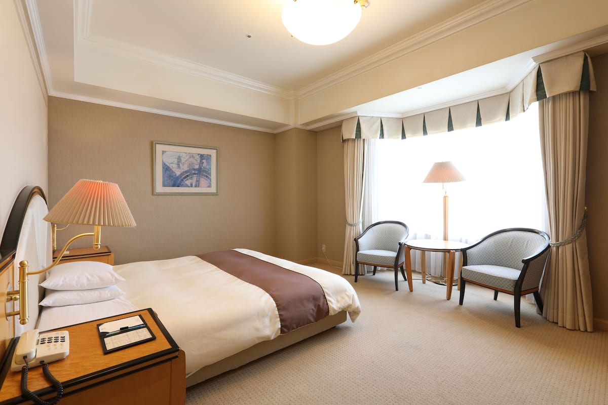 A hotel room with a bed, bedside table with a lamp, a chair, a small table, and a window with curtains.