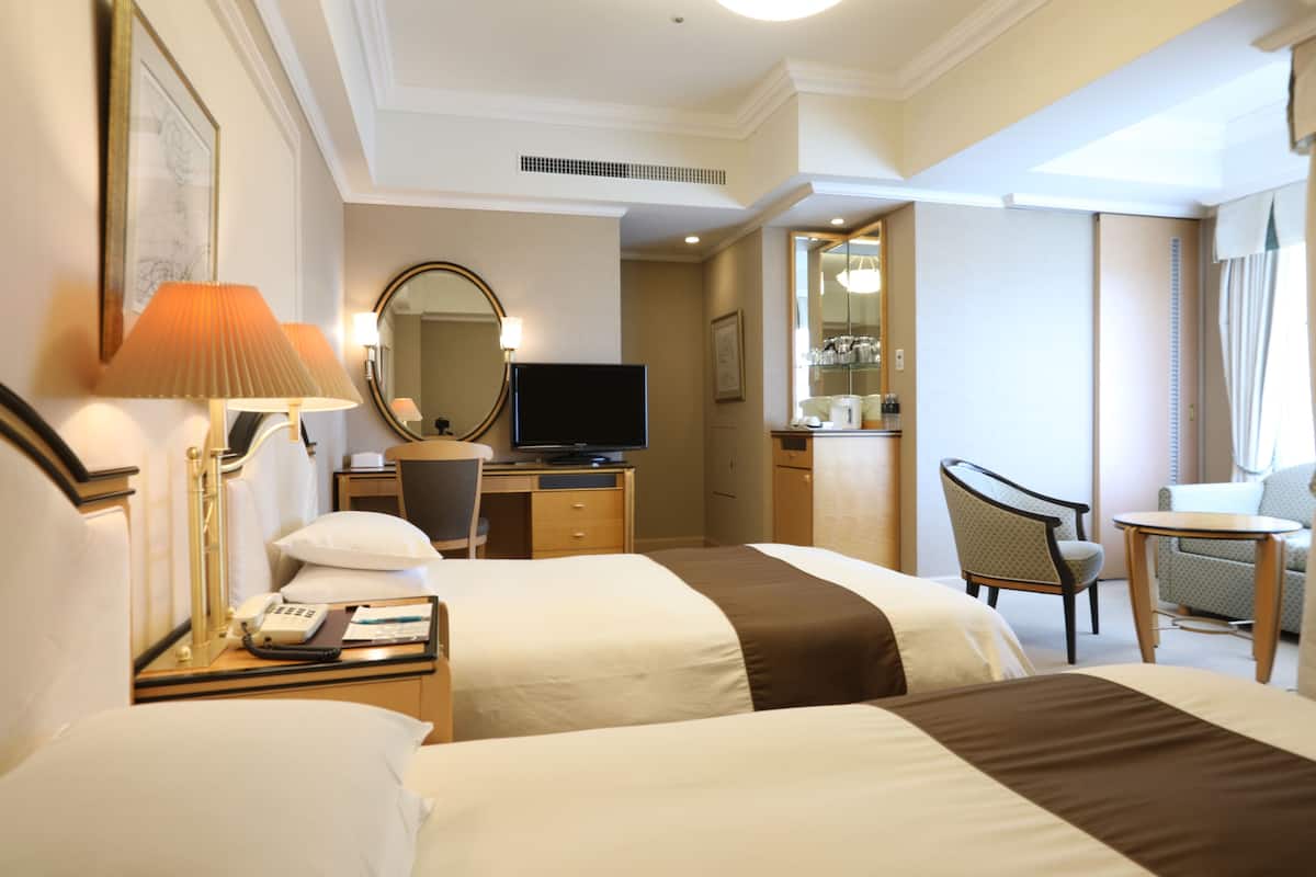 A hotel room with two beds, a desk with a TV, a chair, a small table, and a window with curtains.