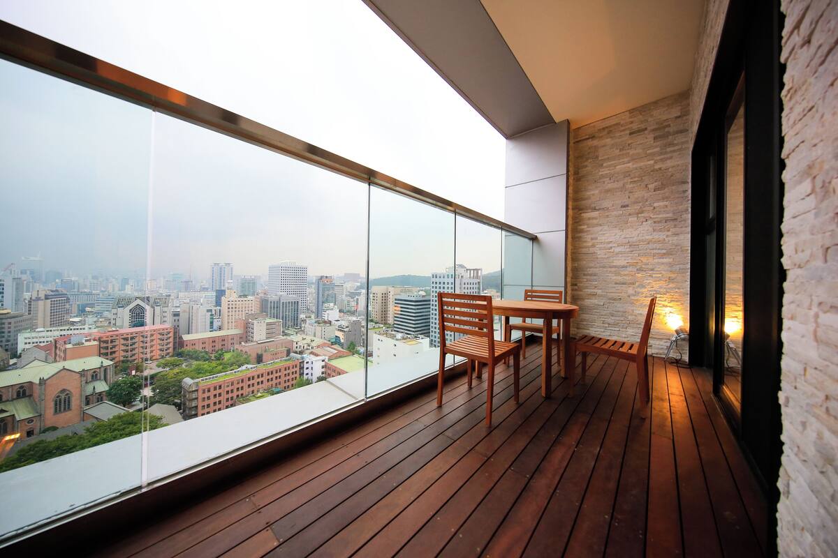 Un balcón con piso de madera, una mesa y sillas. Se aprecia una vista de la ciudad con edificios e iglesia.