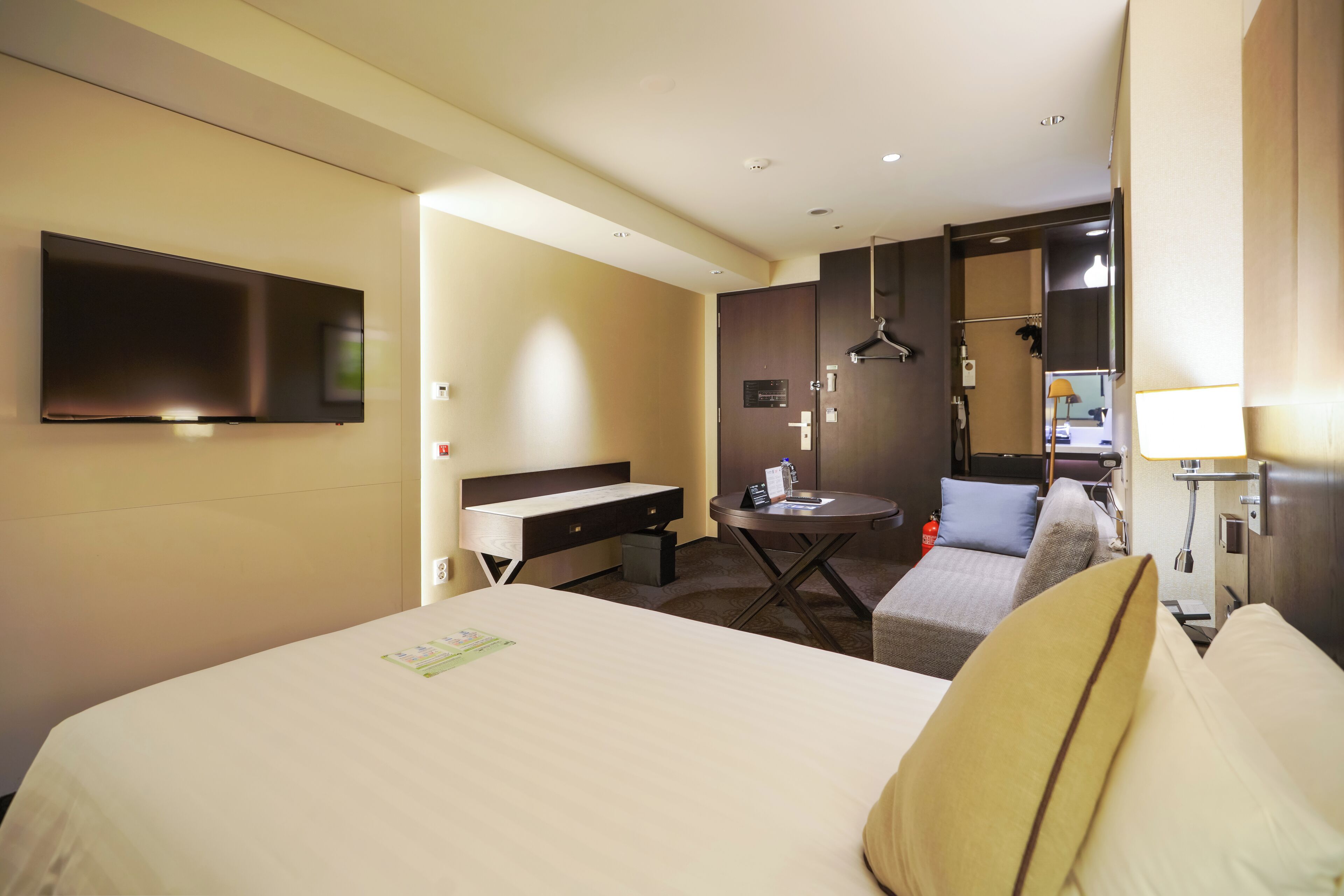 A hotel room with a bed, a desk, a chair, a small table, a TV, and a bathroom area.