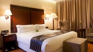 Habitación de hotel con una cama grande, dos lámparas de noche, una mesita de noche, una silla y una pequeña mesa con un jarrón.