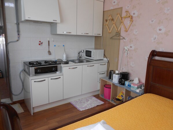 Une cuisine compacte avec des meubles blancs, un four à micro-ondes, une cuisinière et un évier. On y trouve une chaise en bois et un lit recouvert d’une couverture jaune.