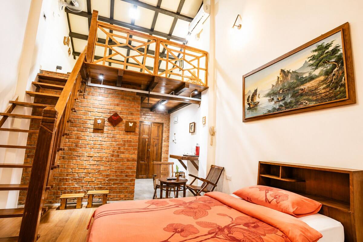 A bedroom with a brick wall, wooden staircase, a bed with orange floral bedding, a wooden headboard, and a painting on the wall.