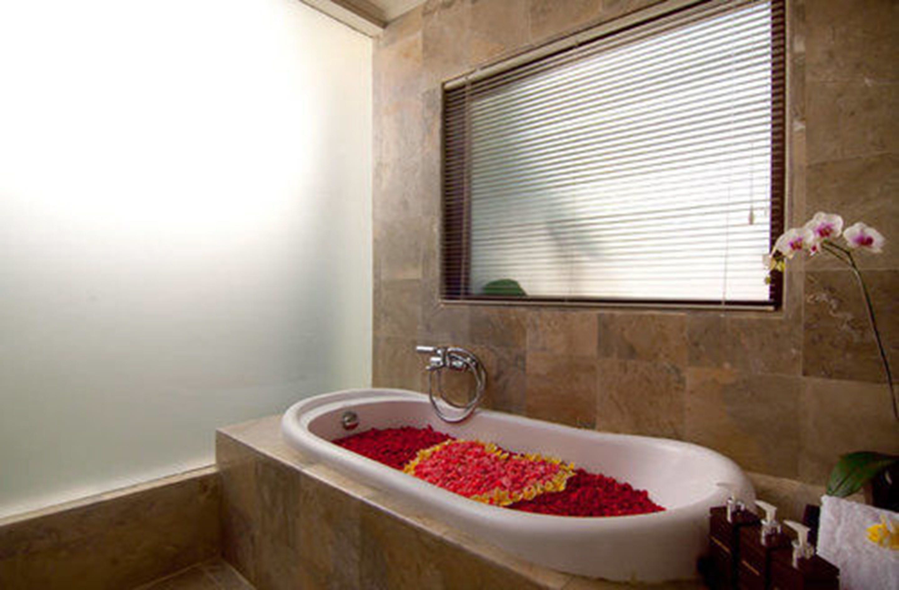 Un bagno con una vasca da bagno piena di petali di rosa, una finestra con persiane e un'orchidea in vaso.