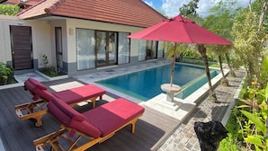 Area piscina con lettini di legno e un ombrellone rosso, una piscina e una casa con tetto in tegole.