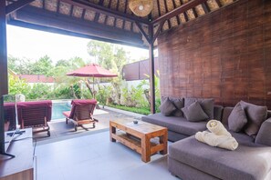 A covered outdoor seating area with a pool, wooden furniture, and a sofa with a towel. - The Awan Villas (Seminyak)