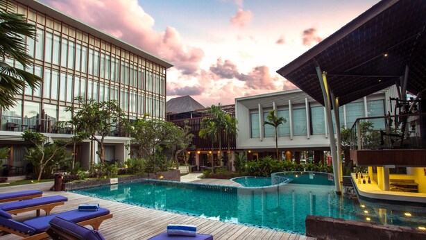 A resort pool area with lounge chairs, a wooden deck, and a modern building with large windows. - The Lerina Hotel Nusa Dua (Nusa Dua)