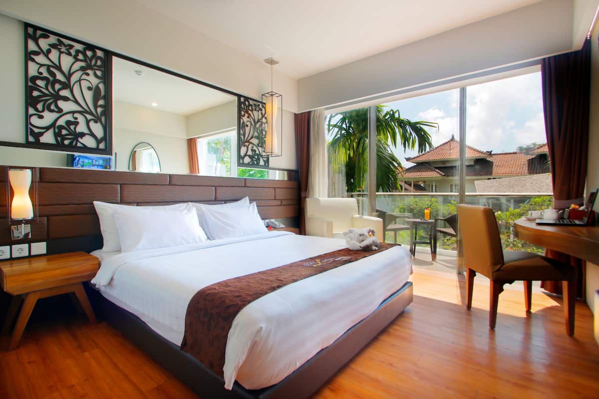 Habitación de hotel con una cama grande, cabecera de madera, escritorio con silla, ventana amplia con vista a palmeras y casas, y un adorno en la pared.