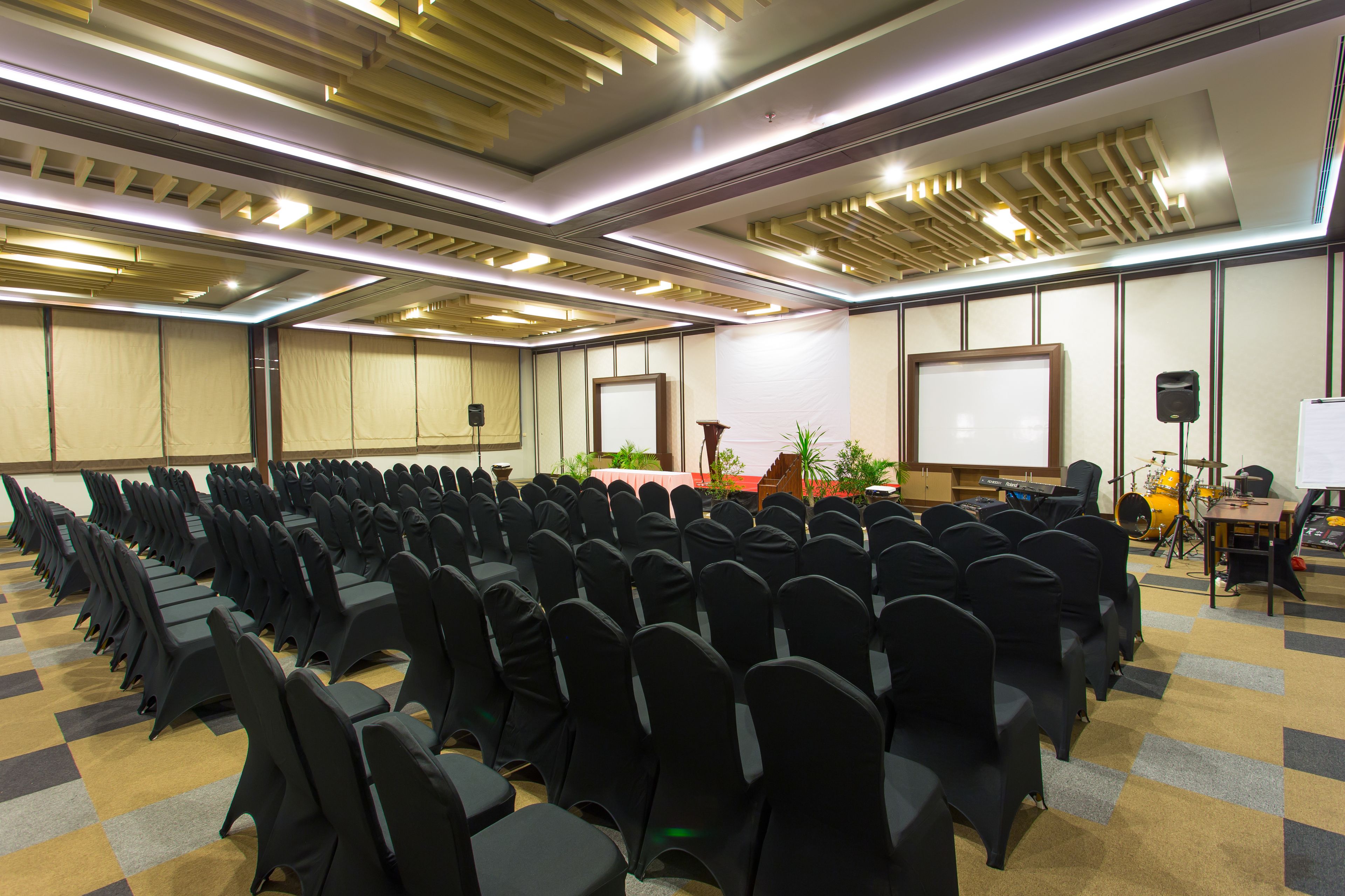 A conference room with rows of chairs, a stage with a podium, and a drum set.