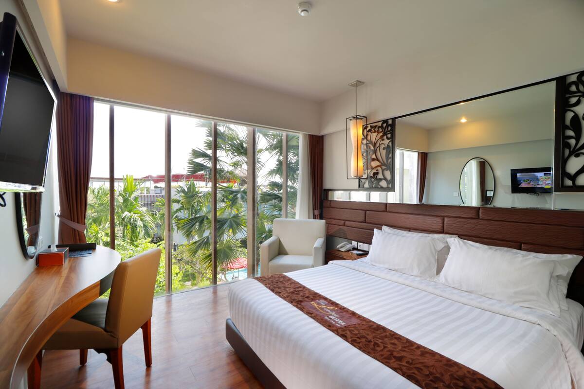Habitación de hotel con una cama grande, un escritorio con silla, televisión y vistas a palmeras a través de amplias ventanas.