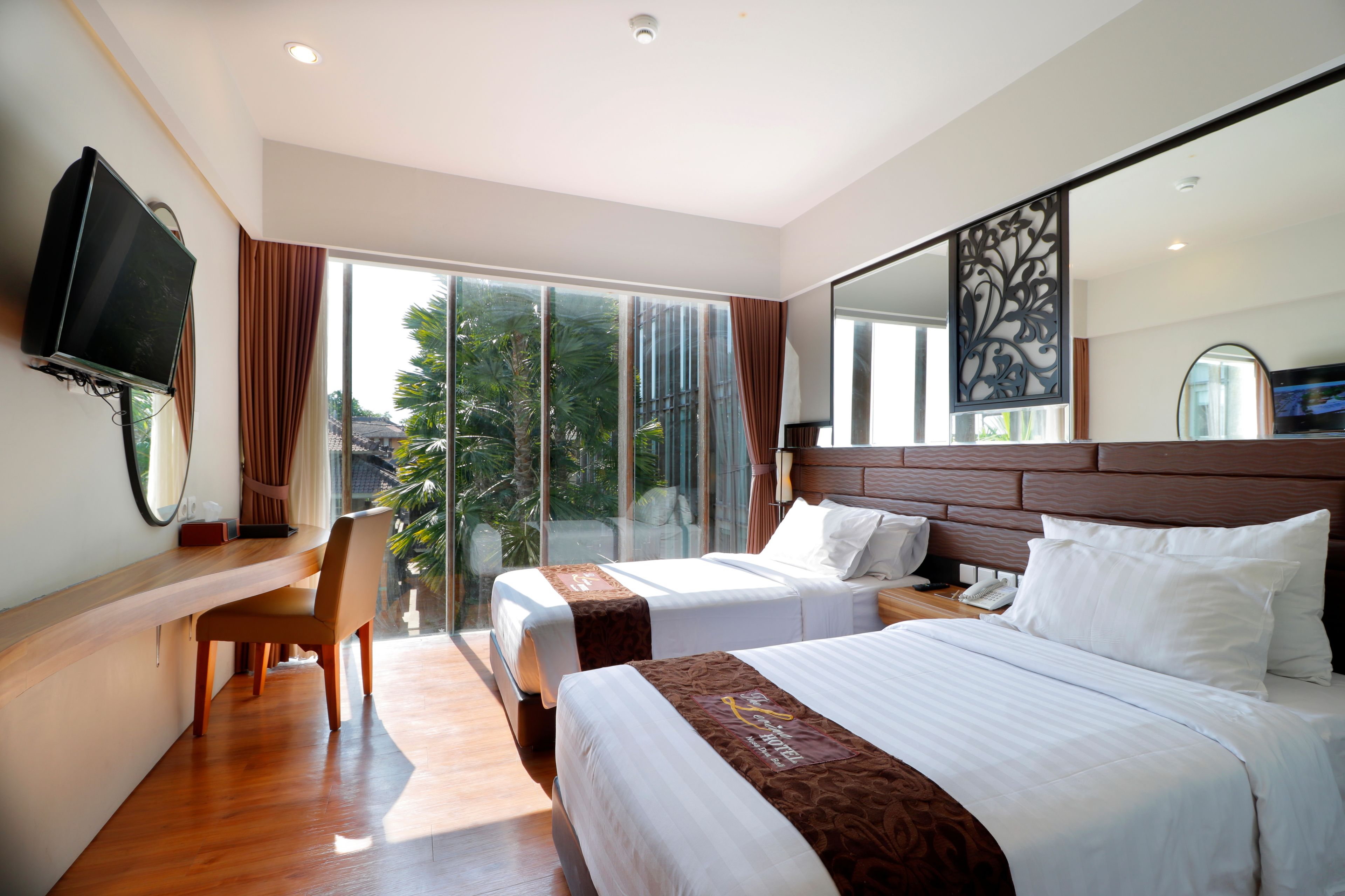 A hotel room with two beds, a desk, a TV, and a large window with a view of greenery.
