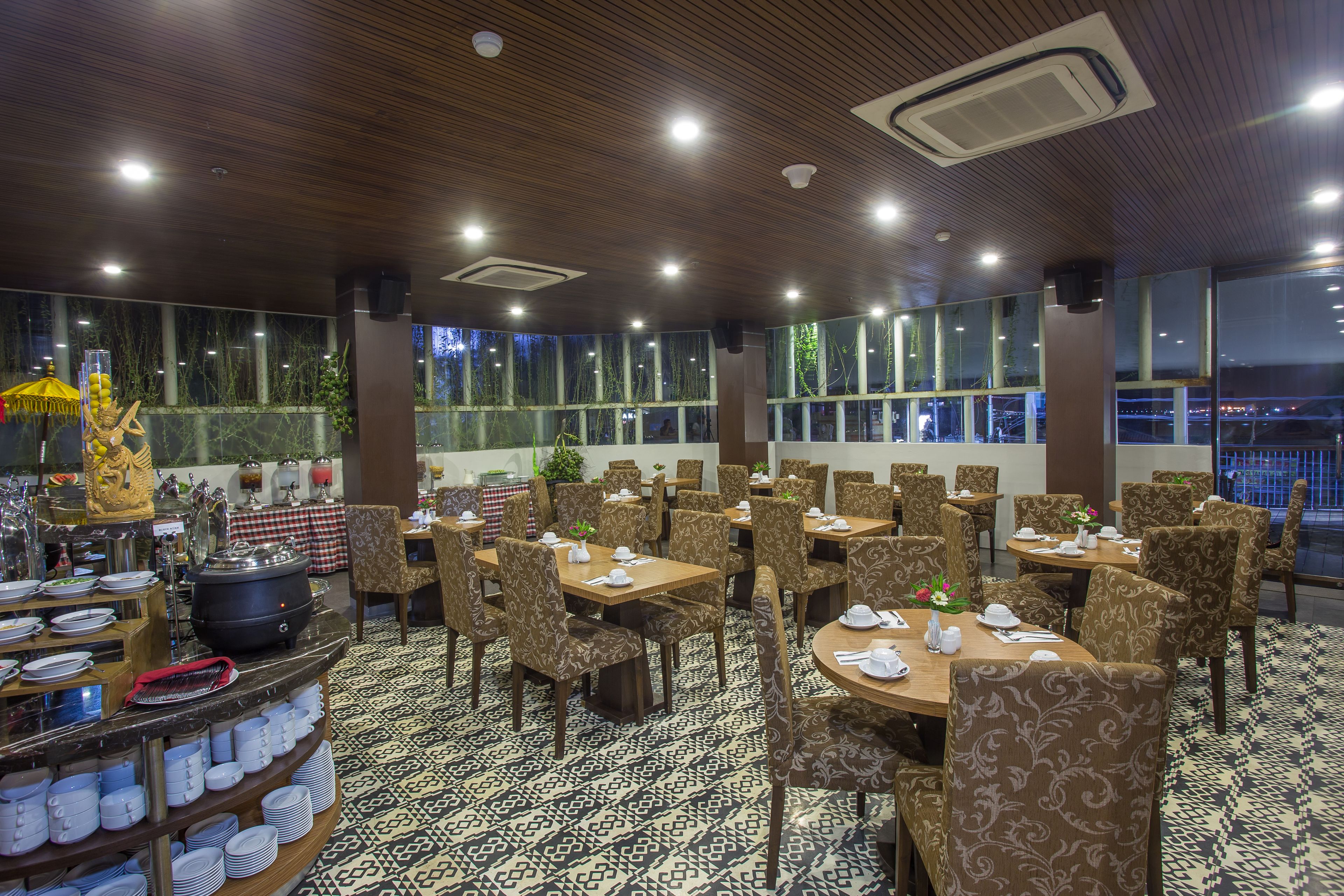 A restaurant with multiple tables and chairs, a buffet area with dishes, and large windows offering a view of the outside.
