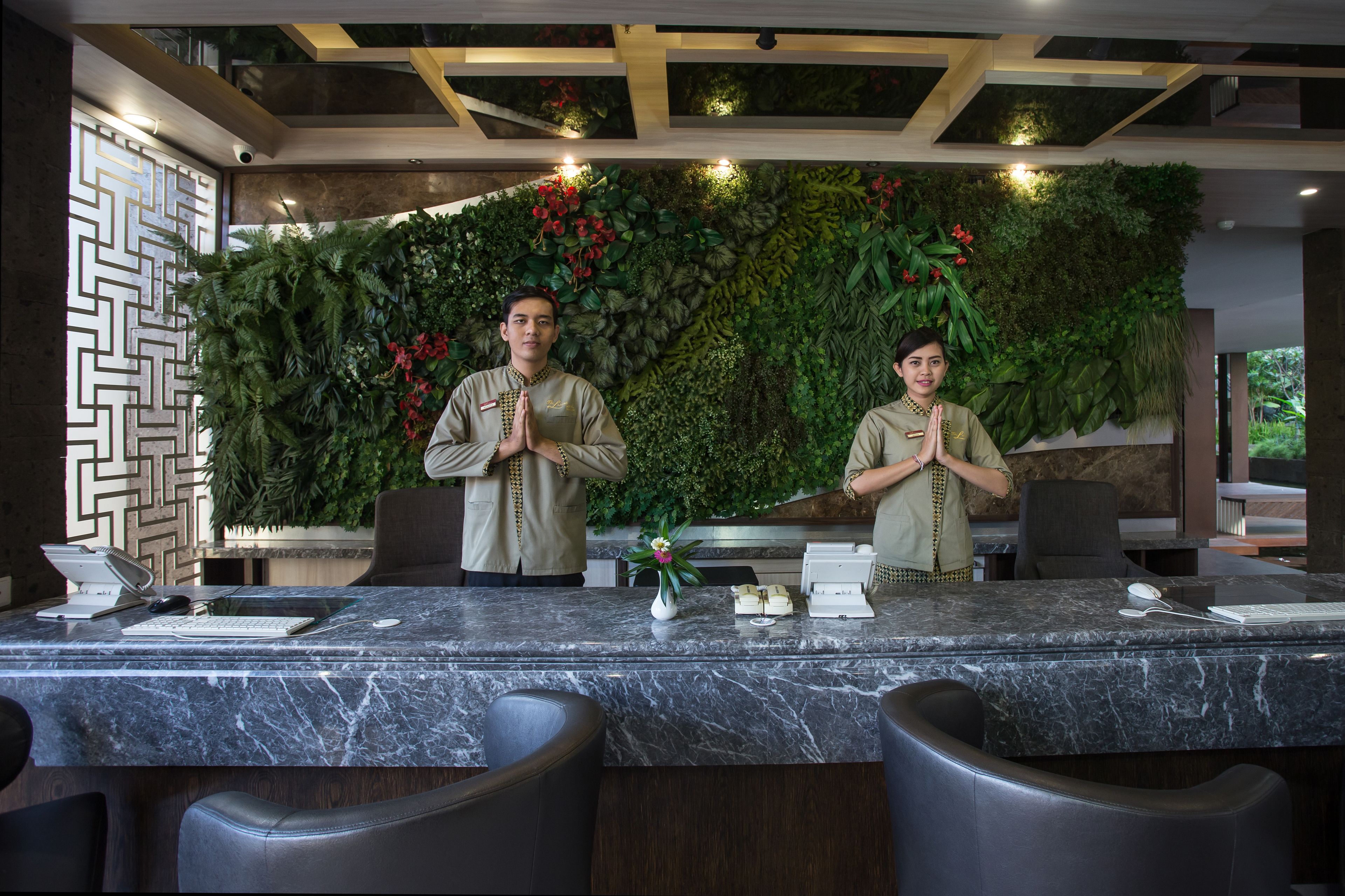 A reception desk with a marble top, two staff members in uniform, a decorative wall with plants, and a geometric wall pattern to the left.