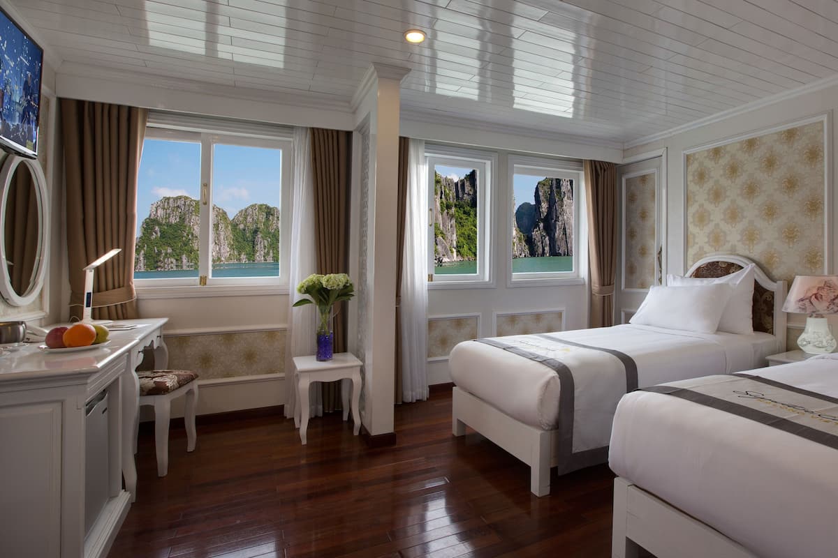 A hotel room with two beds, a desk with a lamp, a chair, and a view of a mountainous landscape through the windows.