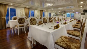 A formal dining room with white tables and chairs, a wooden floor, and large windows with curtains. - Signature Royal Halong Cruise (Hong Gai)