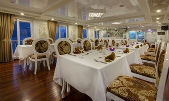Un comedor formal con mesas y sillas blancas, piso de madera y ventanas grandes con cortinas.