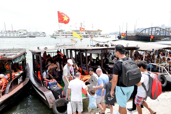 A crowded boat with passengers and crew, docked at a port with other boats and a bridge in the background.