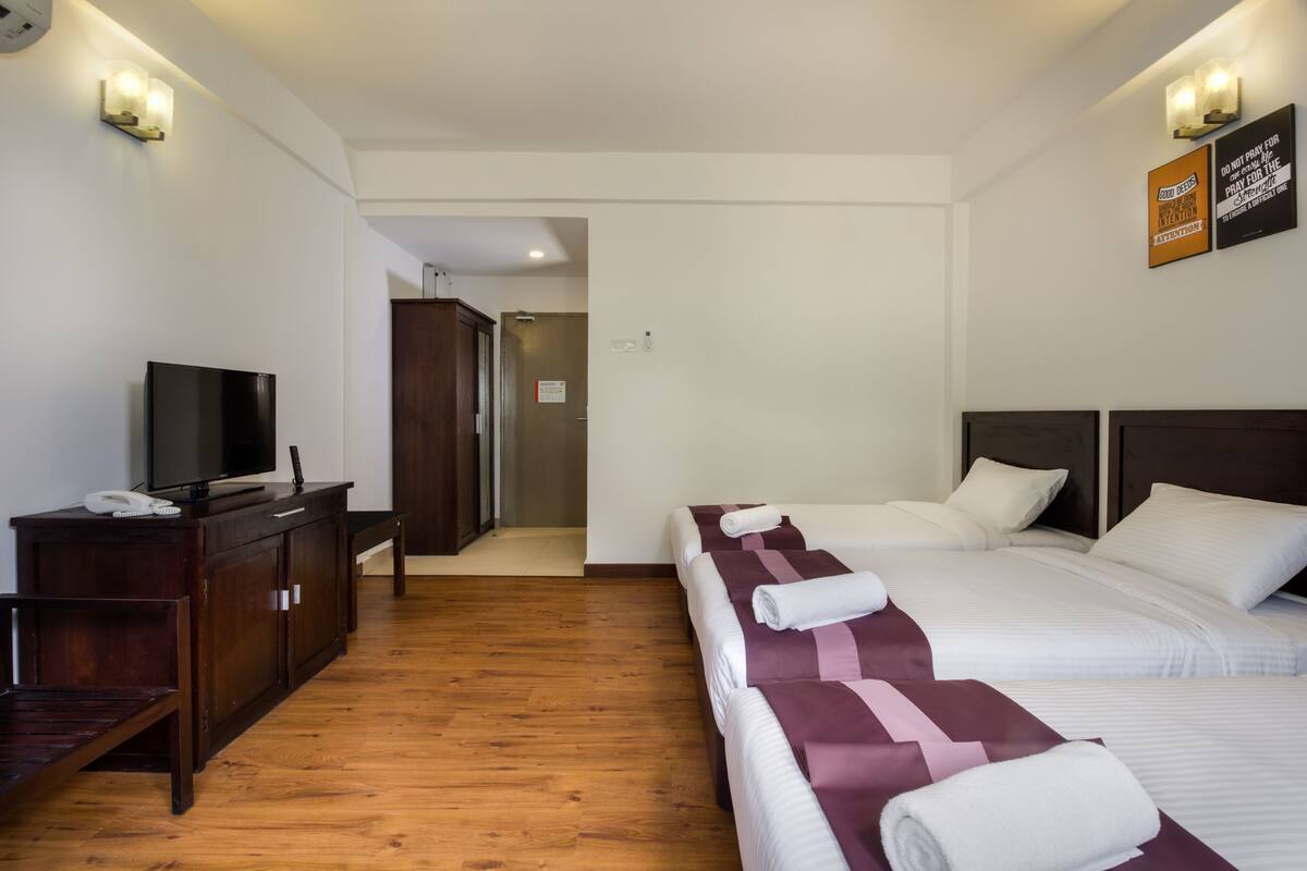 Quarto de hotel com piso de madeira, televisão sobre um móvel de madeira, duas camas com roupa de cama branca e roxa, e luminária fixada na parede.