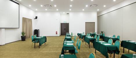 Un salón de conferencias con filas de mesas, sillas y una pantalla de proyector.