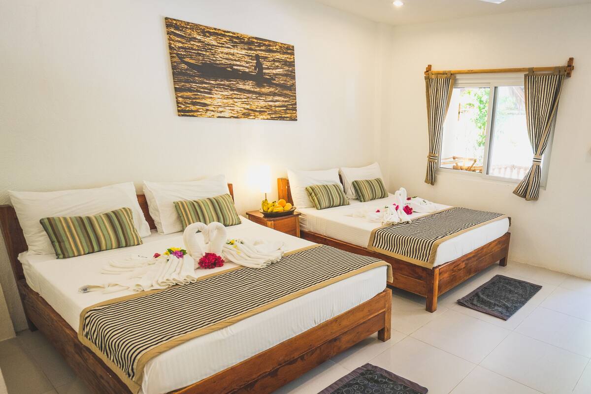 A hotel room with two wooden beds, a painting of a sunset over water, a window with curtains, and a nightstand with a lamp and fruit.