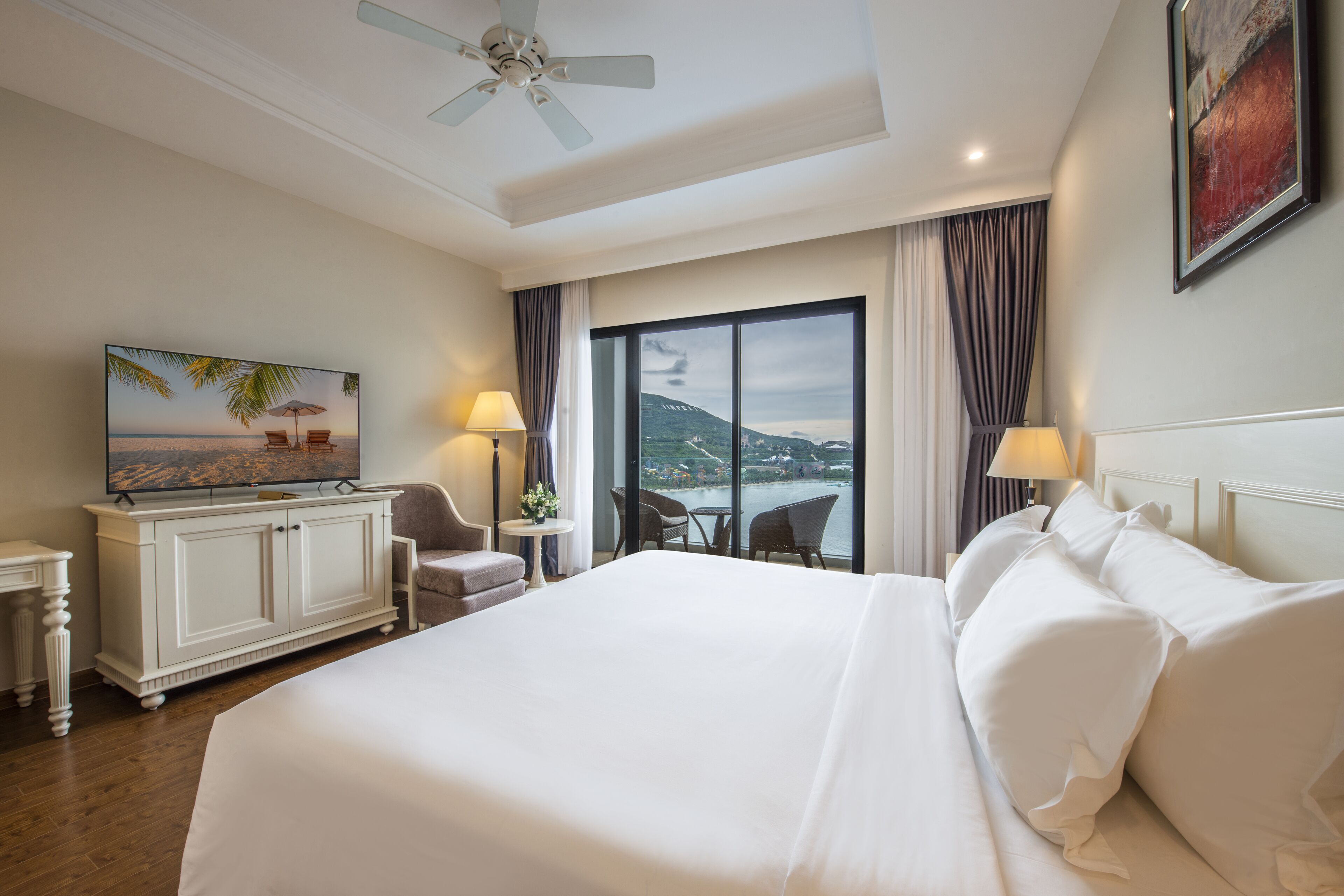 Una camera d'albergo con un letto grande, una televisione su un mobile, una scrivania con una lampada e vista sul mare dalla finestra.