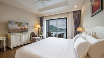 Un cuarto de hotel con una cama grande, un televisor sobre un mueble, un escritorio con lámpara y vista al mar por la ventana.