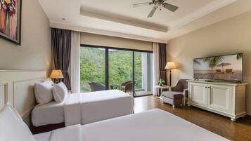 Habitación de hotel con dos camas, un escritorio con televisor, una silla y vista a un bosque a través de ventanas amplias.