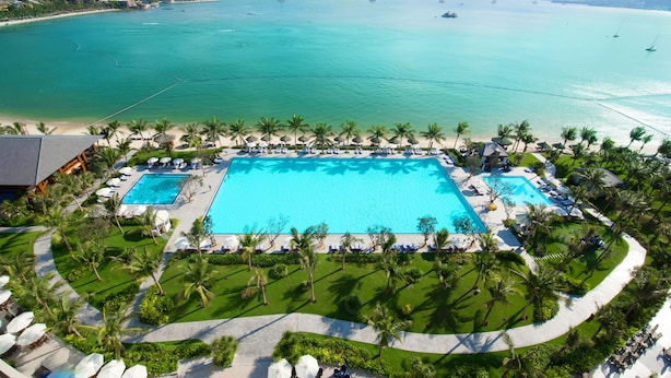 Un resort con una amplia piscina central, rodeado de palmeras y vistas a la playa.