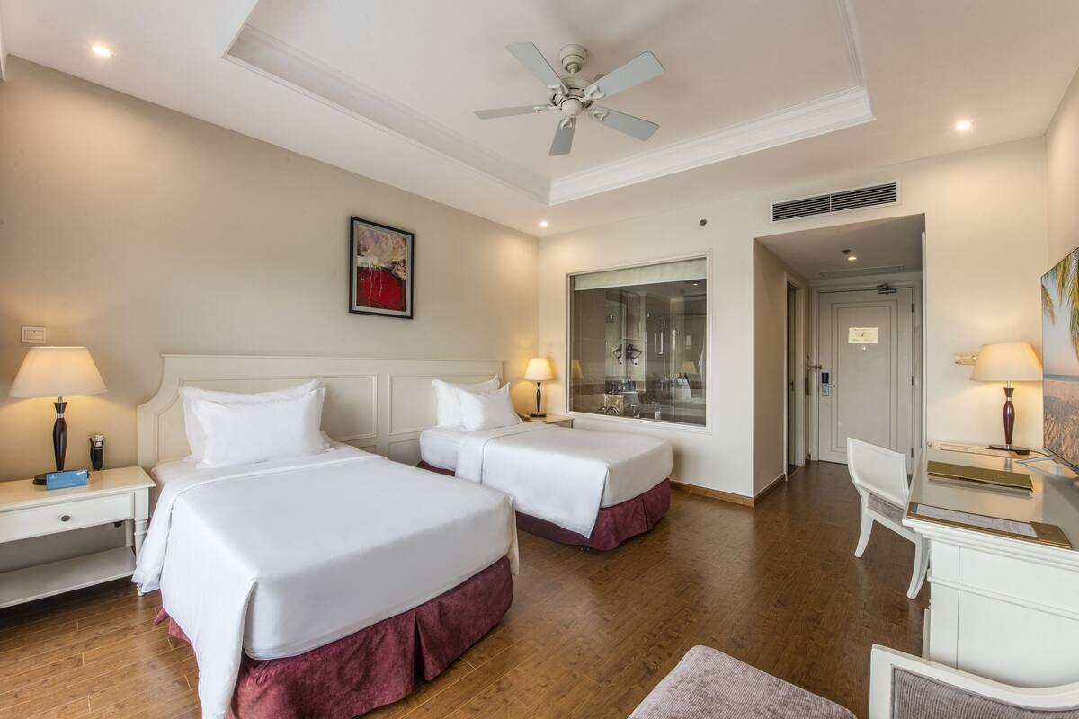 A hotel room with two beds, a desk, a chair, a ceiling fan, and a large window.