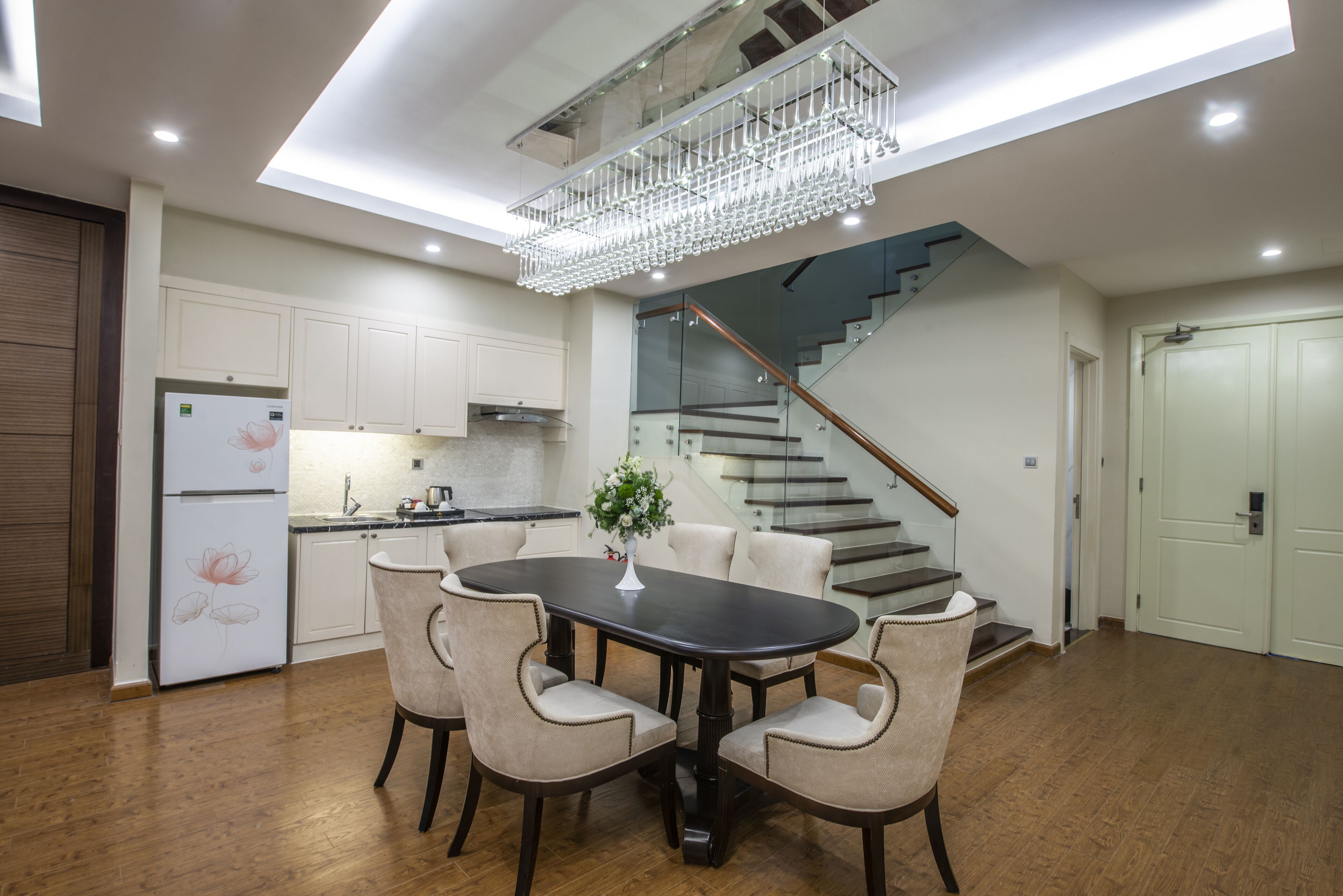 A modern kitchen with white cabinets, a stainless steel refrigerator, and a dining area with a dark wooden table and beige chairs.