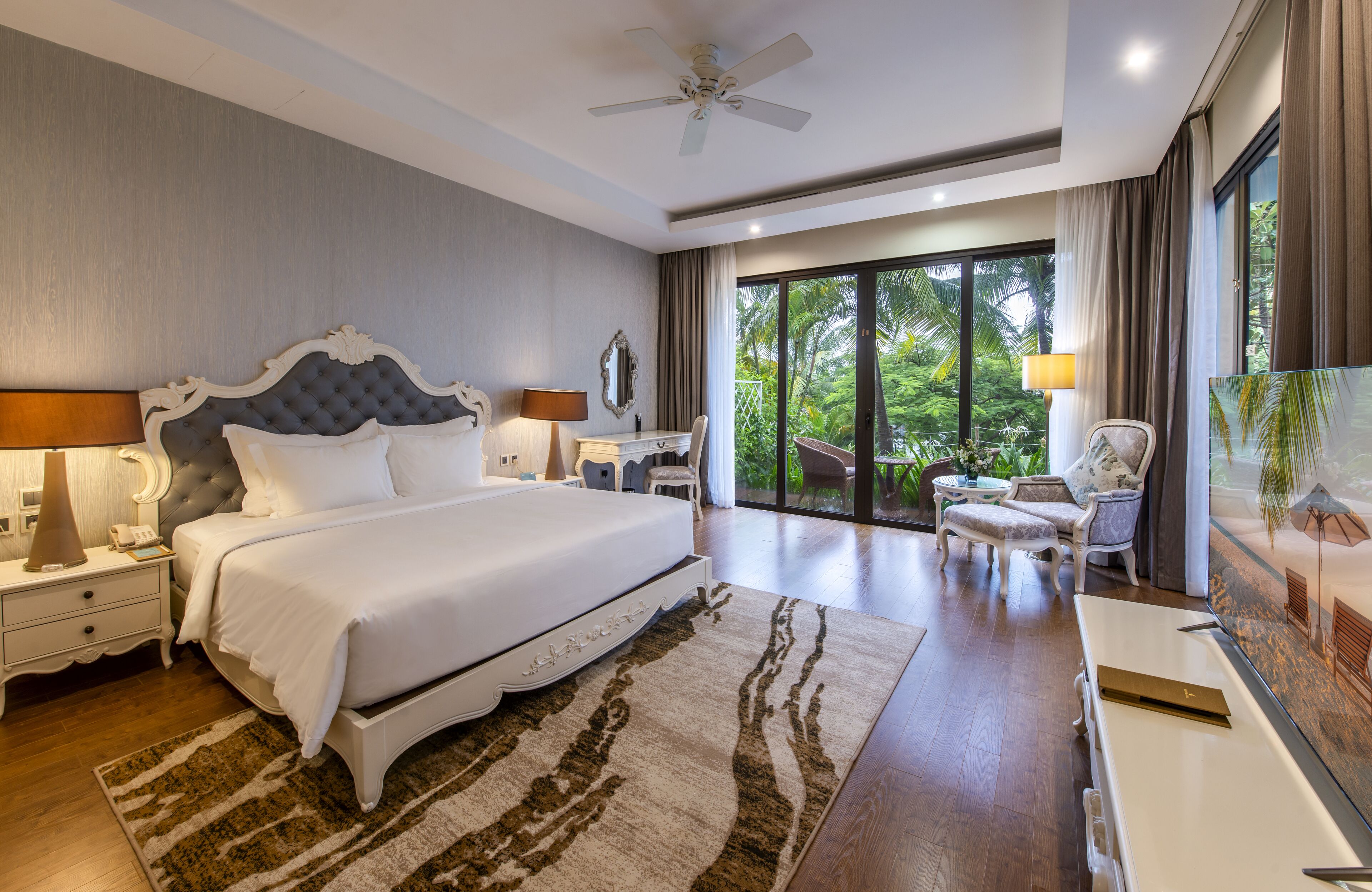A spacious bedroom with a large bed, a ceiling fan, a desk, a chair, and a large window offering a view of the outdoors.