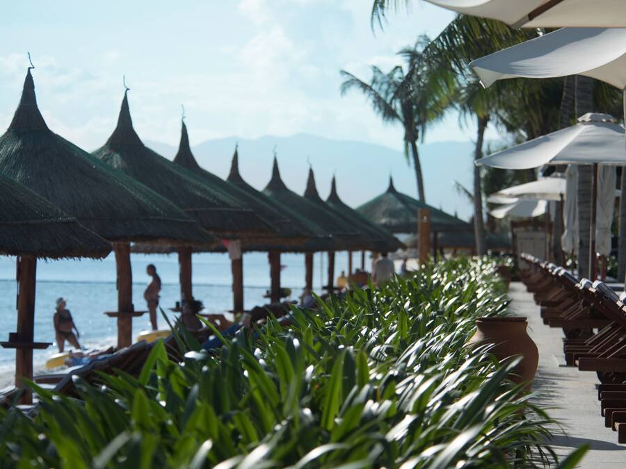 Un complexe hôtelier en bord de mer, avec de nombreuses rangées de parasols en chaume et de chaises de plage, offrant une vue sur l’océan.