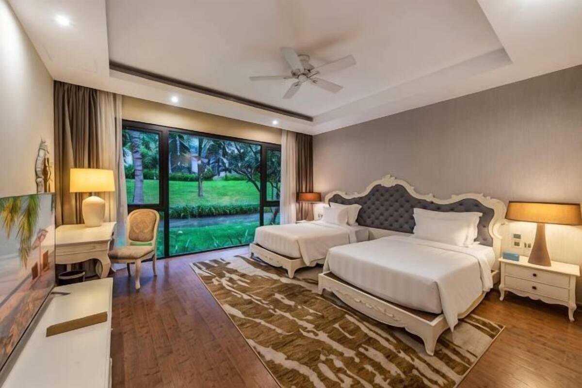 A bedroom with two beds, a desk, a chair, and a view of a garden through large windows.