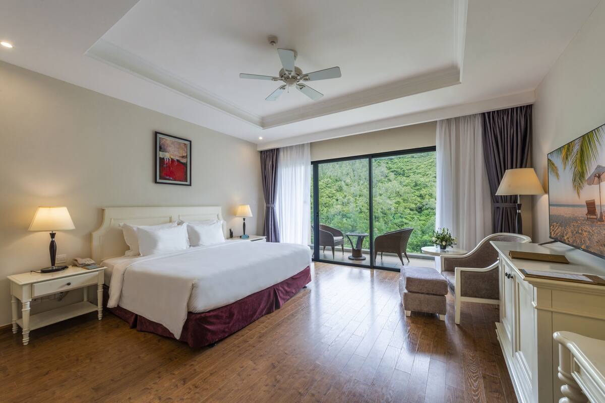 A hotel room with a large bed, two bedside tables with lamps, a ceiling fan, a balcony with a table and chairs, and a view of greenery outside.