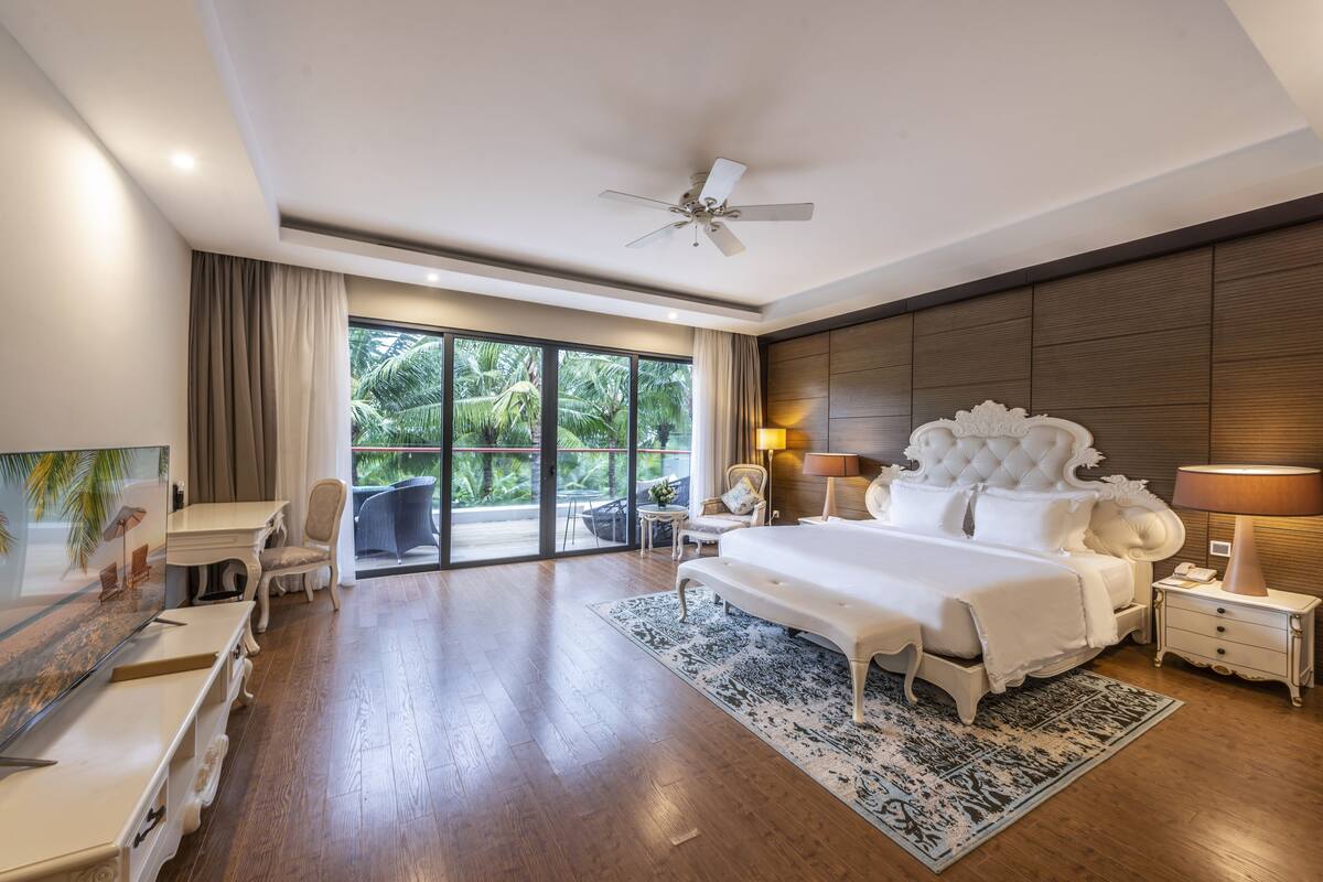 A spacious bedroom with a large bed, a ceiling fan, a desk, and a view of the outdoors.
