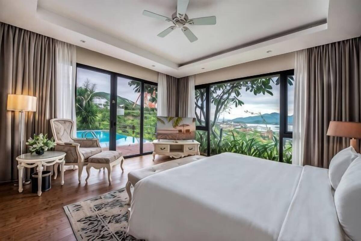 A bedroom with a bed, a desk, a chair, a TV, and a view of a swimming pool and mountains.