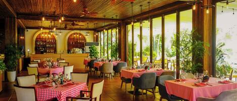 A restaurant with red and white checkered tablecloths, wooden ceiling, and large windows with plants.