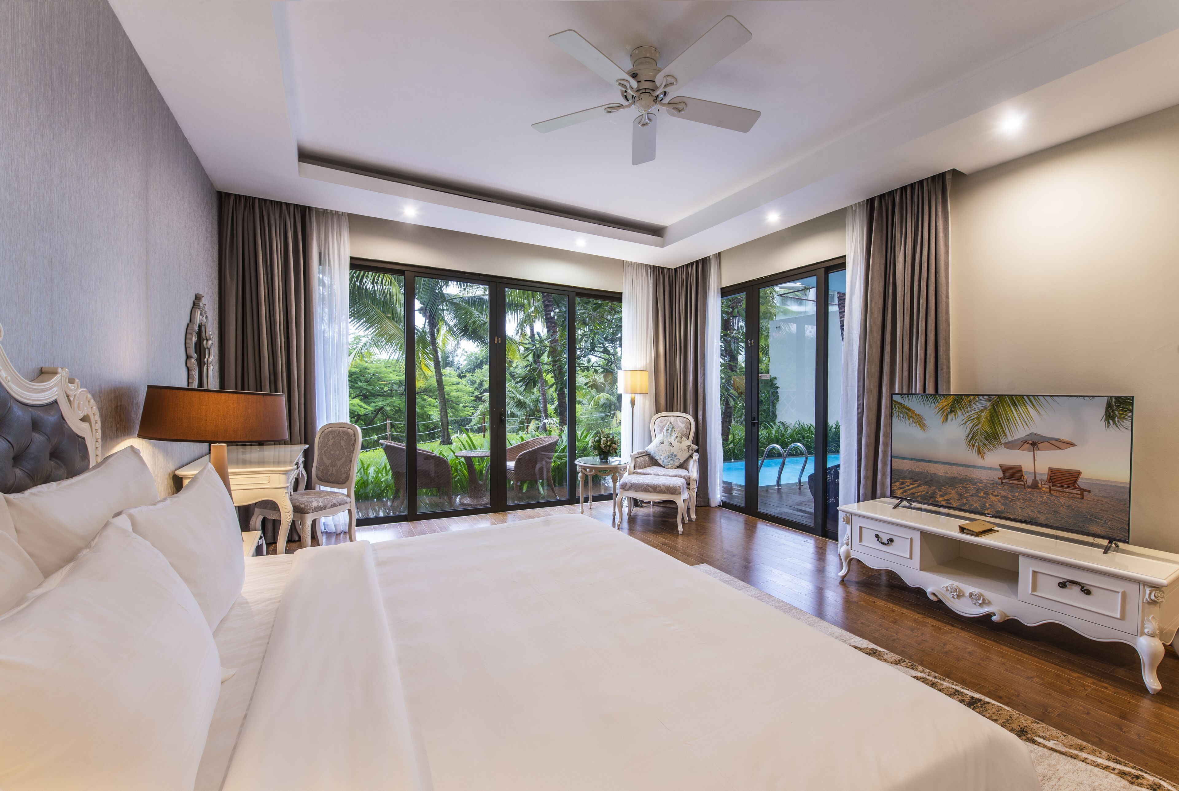 A spacious bedroom with a large bed, a ceiling fan, a TV, and a view of a swimming pool.