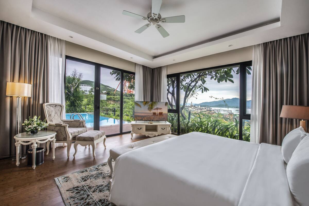 A spacious bedroom with a large bed, a desk, a chair, and a view of a swimming pool and mountains.