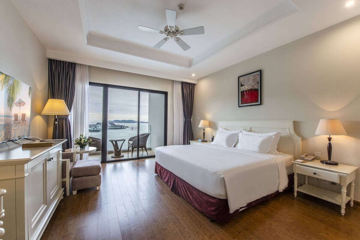 A hotel room with a bed, bedside tables, a desk, and a view of the sea.